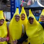A group of people dressed in bright yellow banana costumes smile and pose together outdoors at an Austin bucket-list marathon event. Flags and tents are visible in the blurred background. Austin Marathon Half Marathon & 5K