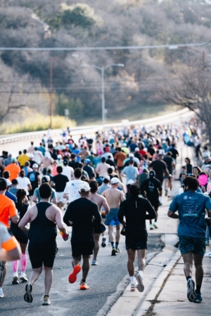 A large group of runners, seen from behind, participates in a road race on a paved street lined with trees—an ideal setting for Austin Marathon hill training as the crowd stretches into the distance under a clear sky. Austin Marathon Half Marathon & 5K