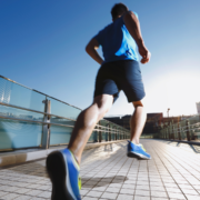 A man in athletic clothes jogs on a city bridge under a clear blue sky, embracing endurance and heat training as the sun shines brightly, with modern buildings in the background. Austin Marathon Half Marathon & 5K