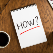 A spiral notebook on a wooden desk displays the word “HOW?” in bold black letters with a red underline. A cup of coffee and part of a keyboard are visible nearby, hinting at February plans for endurance or heat training sessions ahead. Austin Marathon Half Marathon & 5K