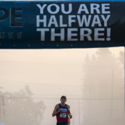 A runner in a race passes under a large banner that reads "YOU ARE HALFWAY THERE!"—a perfect scene for a February Goal or Mid-Year Fitness Check, with fog and trees visible in the background. Austin Marathon Half Marathon & 5K
