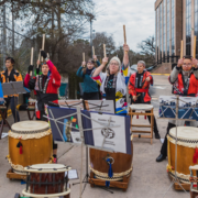 A group of people dressed in colorful traditional attire play large Japanese drums outdoors, each holding drumsticks raised mid-performance—an Austin bucket-list marathon of rhythm. Sheet music stands are visible, with trees and a building in the background. Austin Marathon Half Marathon & 5K