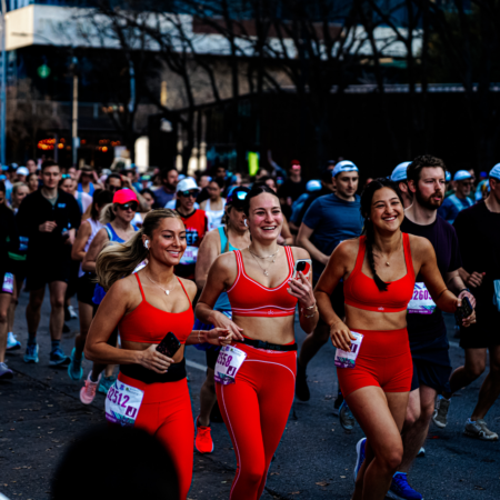 Three women in matching red athletic outfits smile and run together during Race Weekend, surrounded by other runners on a city street. The atmosphere is energetic and lively—perfect for an unforgettable Austin vacation. Austin Marathon Half Marathon & 5K