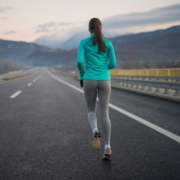 A woman wearing a teal jacket and gray leggings runs alone on an empty highway with mountain scenery in the background under a cloudy sky, staying focused on her February goal. Austin Marathon Half Marathon & 5K