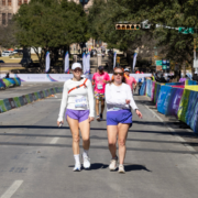 Two women wearing white tops and purple shorts walk side by side on a sunny street during a race event, showcasing their endurance as they navigate colorful barriers and other participants in the background. Austin Marathon Half Marathon & 5K