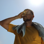 A man in a yellow shirt with a towel around his neck shields his eyes from the bright February sun with his hand, standing outdoors under a clear, blue sky during an endurance heat training session. Austin Marathon Half Marathon & 5K