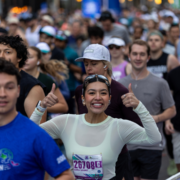 A smiling woman in a white shirt gives two thumbs up while running the Austin bucket-list marathon, surrounded by other energetic runners in athletic gear. She wears a race bib labeled “26708G” and radiates happiness and energy. Austin Marathon Half Marathon & 5K