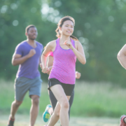 A woman in a pink tank top runs outdoors with two others, all appearing to enjoy heat training on a sunny day, surrounded by lush greenery. Austin Marathon Half Marathon & 5K