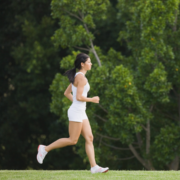 A woman in white athletic wear jogs on grass, with lush green trees in the background—embracing the Summer Consistency Challenge to stay on track for 30 days. Austin Marathon Half Marathon & 5K