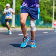 A runner with a bandaged knee jogs on a paved road during a race, showing determination from training. He wears blue running shoes and shorts, while other runners and greenery appear in the blurred background. Austin Marathon Half Marathon & 5K