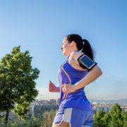 A woman in sportswear jogs outdoors on a sunny day, following her half marathon training plan. She wears earphones and an armband with a phone, with trees and a cityscape behind her under a clear blue sky. Austin Marathon Half Marathon & 5K