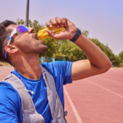 A man in sporty attire and sunglasses is on a running track, squeezing and drinking from a yellow energy gel pack—a perfect snapshot of marathon training. Trees and a clear sky are visible in the background. Austin Marathon Half Marathon & 5K