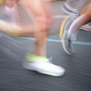 Blurred close-up of runners' legs and feet in motion on a paved road, capturing the speed and energy of marathon training. One runner wears white shoes with neon accents. Austin Marathon Half Marathon & 5K