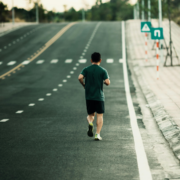 A man wearing a green shirt and black shorts jogs alone on an empty road, heading uphill—perhaps deep into his marathon training. Road signs and trees line the background, setting the scene for focused preparation. Austin Marathon Half Marathon & 5K