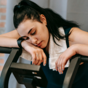 A woman with dark hair and a towel around her neck leans tiredly on a treadmill, looking down with a fatigued expression—a reminder of the importance of burnout prevention during intense training. Austin Marathon Half Marathon & 5K