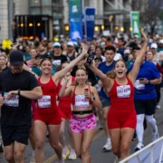 A large group of runners participating in the Austin Marathon; three women in red athletic wear at the front are smiling, raising their arms, and holding hands as they run, with other runners and urban buildings in the background—a true marathon countdown moment. Austin Marathon Half Marathon & 5K