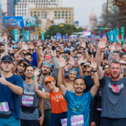 A large, diverse group of marathon runners smile and wave at the camera before a race, sporting colorful athletic gear and numbered bibs. Urban buildings and a capitol building rise behind them, celebrating months of marathon training together. Austin Marathon Half Marathon & 5K