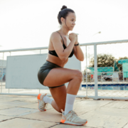 A woman wearing a sports bra, shorts, and sneakers performs a lunge exercise outdoors on a tiled surface near a fence, staying active as her Austin Marathon countdown continues. Buildings and trees are visible in the background. Austin Marathon Half Marathon & 5K