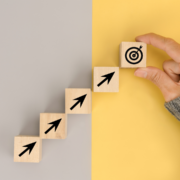 A hand places a wooden block with a target icon at the top of a staircase made of blocks with upward-pointing arrows, symbolizing the 24 weeks countdown to the Austin Marathon, set against a split gray and yellow background. Austin Marathon Half Marathon & 5K