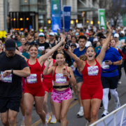 A group of runners participate in the Austin Marathon, with three women in the front smiling and raising their arms in celebration. Many participants are visible behind them, running on a city street and sharing in the exhilarating race experience. Austin Marathon Half Marathon & 5K