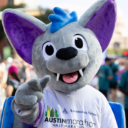 A person in a gray bat costume with large purple ears and blue accents points toward the camera, wearing an Austin Marathon Half Marathon T-shirt at an outdoor February event, ready to commit to the race season ahead. Austin Marathon Half Marathon & 5K