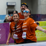 Three smiling runners in orange shirts pose at a race finish line, wearing sunglasses and race bibs. One gives a thumbs-up. Their shirts read, “I THOUGHT THIS WAS A BAR CRAWL?” Colorful event banners are in the background, capturing the lively spirit of a summer run and celebrating their commitment to finishing strong. Austin Marathon Half Marathon & 5K
