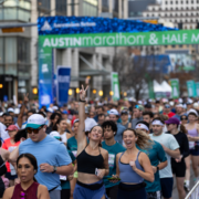 A large crowd of runners smiles and cheers at the starting line of the Austin Marathon & Half Marathon in February, with a green event banner overhead and city buildings in the background, all ready to commit to an inspiring race journey. Austin Marathon Half Marathon & 5K