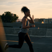 A woman jogs on a city street at sunset, wearing athletic clothes and earphones—embracing February planning for a balanced lifestyle—with buildings and trees visible in the background. Austin Marathon Half Marathon & 5K