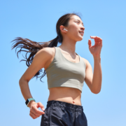 A woman in athletic wear jogs outdoors under a clear blue sky, wearing wireless earbuds and a smartwatch, with her hair in a ponytail—demonstrating how to run in high heat while staying mindful of running safety tips. Austin Marathon Half Marathon & 5K
