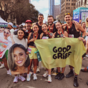 A group of smiling runners pose together after a race, holding medals, a “GOOD GRIEF” flag, and a large cutout of a woman’s face. Tall buildings and other runners are visible in the background, capturing the energy and camaraderie that follow good time management and careful February planning. Austin Marathon Half Marathon & 5K