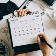 A person holds a pen and a spiral-bound desk calendar open to February 2027, ready for some focused February planning. Office supplies, scissors, and a plate are on the table, creating an ideal workspace for time management and organization. Austin Marathon Half Marathon & 5K