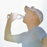 A person wearing a cap and sunglasses drinks water from a plastic bottle outdoors, with sunlight shining brightly in the background—demonstrating important running safety tips to train safely in heat. Austin Marathon Half Marathon & 5K