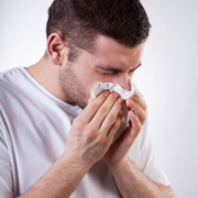 A man in a white shirt is holding a tissue to his nose and appears to be sneezing or blowing his nose, highlighting the importance of running safety tips—especially when you train safely in heat, as allergies and heat can both impact your performance. Austin Marathon Half Marathon & 5K