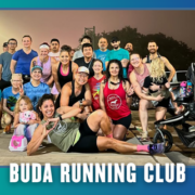 A diverse group of people, including kids and a dog, pose smiling in athletic wear on a sidewalk at dawn with streetlights, ready for their South Austin group run. The text below reads “BUDA RUNNING CLUB.”. Austin Marathon Half Marathon & 5K