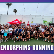 A large group of people pose outdoors in athletic wear under string lights, with surfboards and a New Balance tent in the background. Text at the bottom reads “ENDORPHINS RUNNING.” This spirited Group Run was part of TEAM FX’s KickOff Week celebration. Austin Marathon Half Marathon & 5K