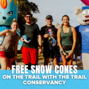 A group of people and two mascots stand smiling outdoors at a fun run event. A blue banner at the bottom reads, “Free snow cones on the trail with the Trail Conservancy,” highlighting this free event for all participants. Austin Marathon Half Marathon & 5K