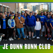 A group of people, some wearing blue “JE Dunn Runn Club” shirts, pose together outdoors near a building in South Austin. A dog is sitting in front. Text at the bottom reads “JE DUNN RUNN CLUB,” highlighting the excitement of kickoff week for their group run. Austin Marathon Half Marathon & 5K