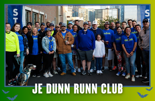 A group of people, some wearing blue “JE Dunn Runn Club” shirts, pose together outdoors near a building in South Austin. A dog is sitting in front. Text at the bottom reads “JE DUNN RUNN CLUB,” highlighting the excitement of kickoff week for their group run. Austin Marathon Half Marathon & 5K