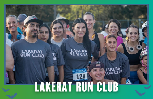 A group of smiling people wearing “Lakerat Run Club” shirts pose together outdoors at a South Austin running event during KickOff Week, with trees in the background and race numbers visible, celebrating their group run. Austin Marathon Half Marathon & 5K