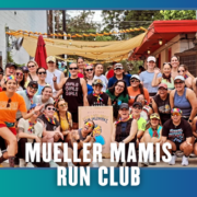 A diverse group of smiling women pose together outdoors, some wearing rainbow accessories and athletic gear, holding a sign that reads “Mueller Mamis Run Club.” Two small dogs are also present, celebrating the kickoff week of their marathon club with this joyful group run. Austin Marathon Half Marathon & 5K