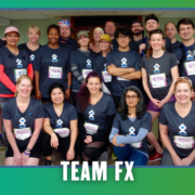 A group of people in matching “TEAM FX” shirts pose indoors, smiling after a Group Run during KickOff Week. They wear race bibs and medals, celebrating their achievement with Goodpain Athletics. The image has a green and blue border with “TEAM FX” written at the bottom. Austin Marathon Half Marathon & 5K