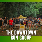 A large group of people poses outdoors at night in running gear in front of a yellow tent and sign, celebrating KickOff Week. Text at the bottom reads, “The Downtown Run Group.” Trees are visible in the background. Austin Marathon Half Marathon & 5K