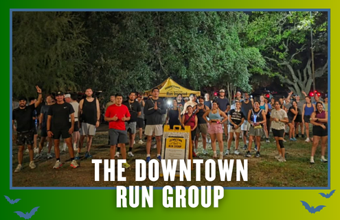 A large group of people poses outdoors at night in running gear in front of a yellow tent and sign, celebrating KickOff Week. Text at the bottom reads, “The Downtown Run Group.” Trees are visible in the background. Austin Marathon Half Marathon & 5K