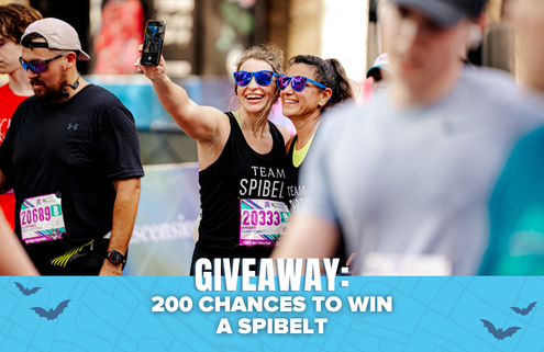 Two women wearing race bibs and sunglasses take a selfie at an outdoor event. Text on the image reads: “GIVEAWAY: 200 CHANCES TO WIN A SPIBELT.” Other participants are visible in the background, highlighting the excitement of the Spibelt giveaway. Austin Marathon Half Marathon & 5K