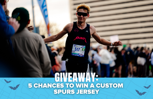 A runner in sunglasses finishes a race with arms outstretched. Text overlay reads: “GIVEAWAY: 5 CHANCES TO WIN A CUSTOM SPURS JERSEY AND YETI COOLER.” Crowd and buildings are visible in the background. Austin Marathon Half Marathon & 5K