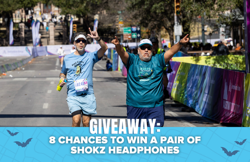 Two runners in blue shirts cheer as they approach the finish line of a race, capturing a moment perfect for a Finisher Pix photo package. A banner at the bottom reads, “GIVEAWAY: 8 CHANCES TO WIN A PAIR OF SHOKZ HEADPHONES.”. Austin Marathon Half Marathon & 5K
