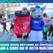 A runner in a strawberry jam costume stands next to a person in a blue wolf mascot suit at the Austin Marathon, sponsored by Ascension Seton, with cheering participants in the background. Text below promotes GOOD GOOD as the official jam and goodie bag. Austin Marathon Half Marathon & 5K